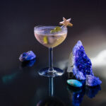 A gin Martini cocktail for Virgo on a tabletop with a star topper in the glass and purple amethyst on the table.