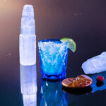 A margarita cocktail for cancer in a small blue glass with a lime garnish and salt rim. Selenite sits near by on the black table.