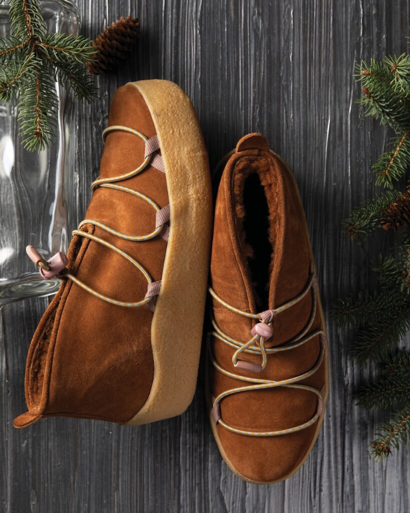 A pair of brown clog boots from a shop in Aspinwall sit on a black textured background between two sprigs of pine.