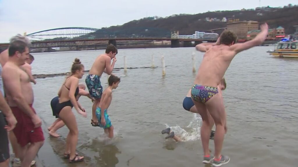 A bunch of men jump into the Monongahela river in Pittsburgh with the bridges in the background.