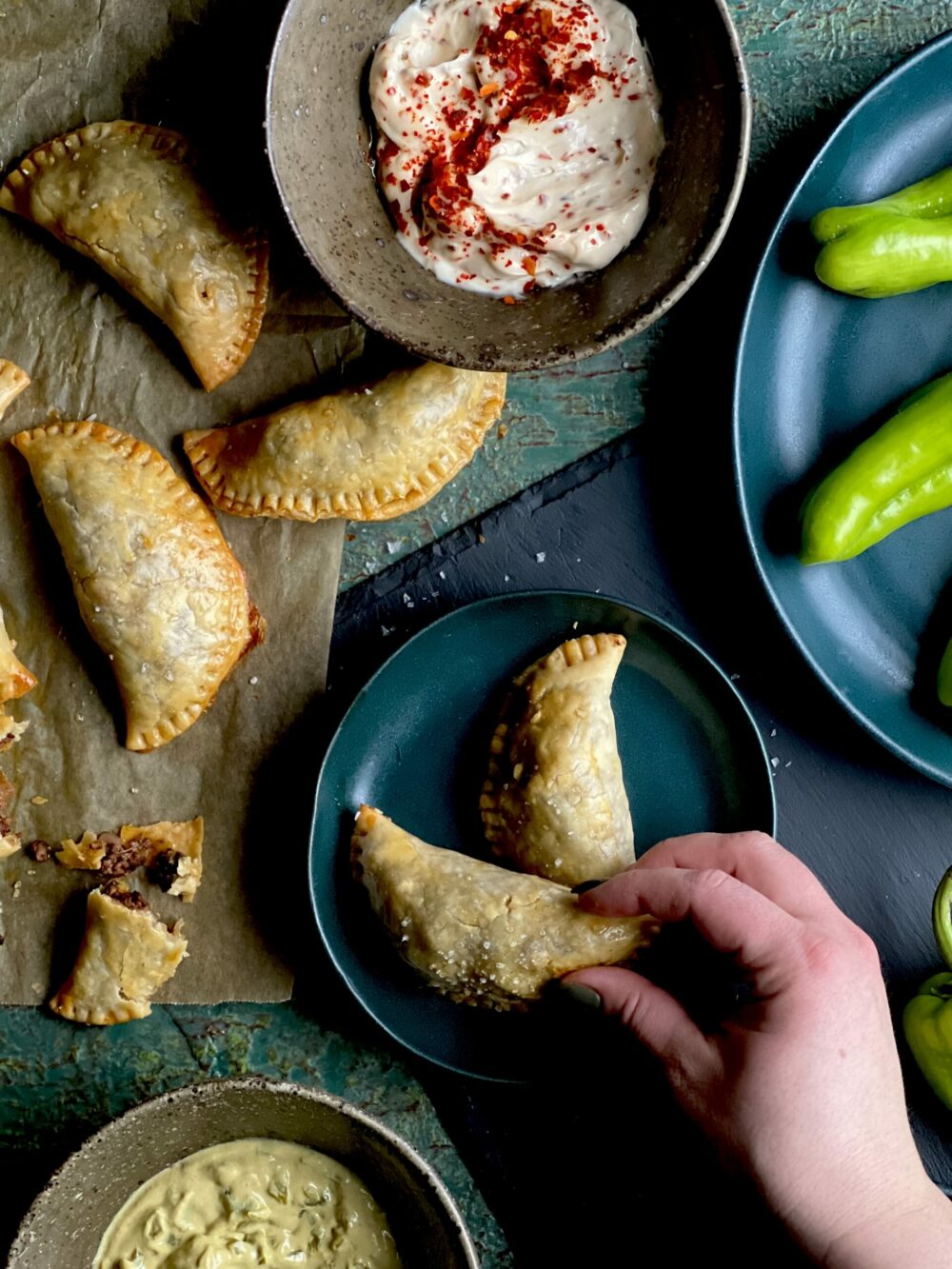 A woman's hand picing a moon shaped hand pie from a small blue dish with and pies dips and raw ingredients surrounding the dish on a brown paper, green and blue surface.