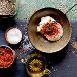 Bowls with seasonings and oil on a black slate surface surrounding a brown bowl of harissa chili mayo dip.