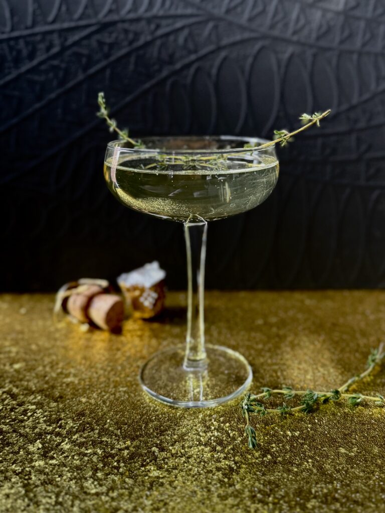 A light colored beverage in a coupe glass with a thyme sprig garnish on a glittery gold surface with a black background.