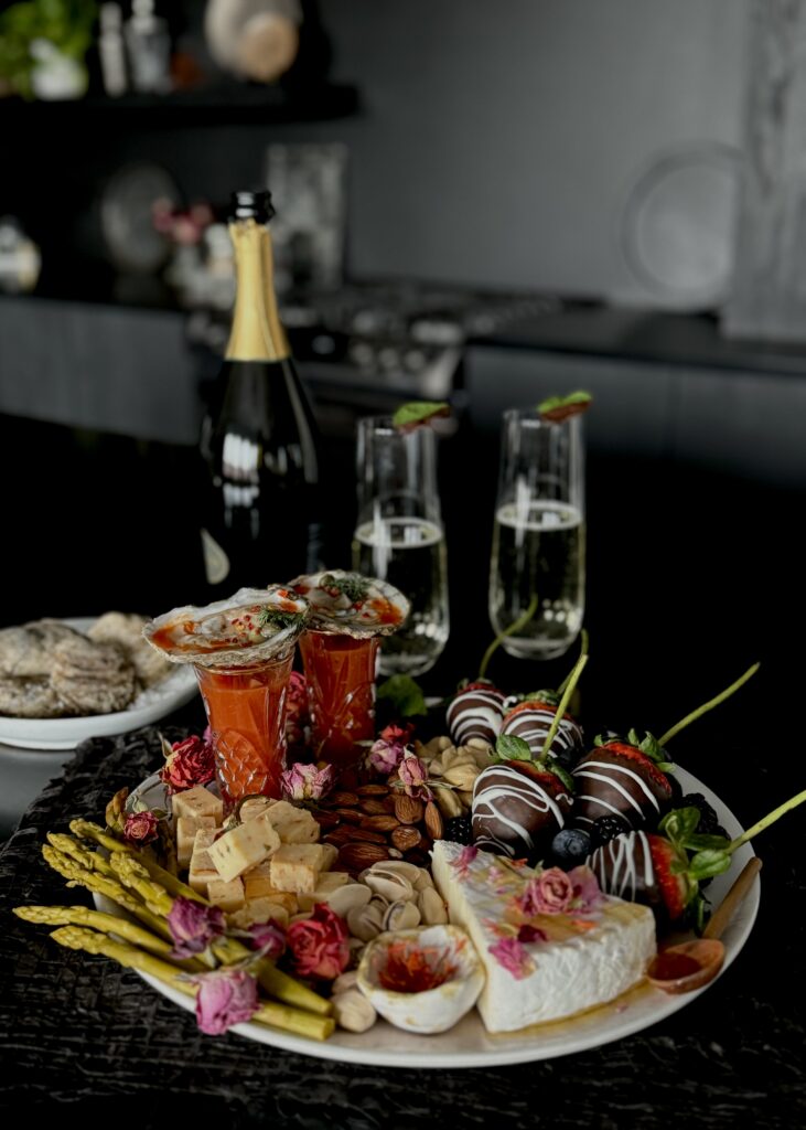 An aphrodisiac board for 2 with several aphrodisiac ingredients like, berries, nuts, cheese, honey, oyster shooters and pickled asparagus on a black counter in a black kitchen with two glasses of champagne, a champagne bottle, and a dish of oysters on the half shell in the background.