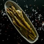 An overhead photo of an oval dish with pickled asparagus spears, a glove of garlic, and a bay leaf, on a black surface with sprinkled salt.