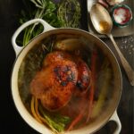 A pot holds an immune boosting bone broth recipe featuring a whole chicken, carrots, and celery. A large spoon and herbs sits beside the pot.