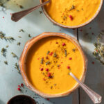 Two soup bowls filled with orange butternut squash soup topped with shredded leaf garnishes and a spoon.