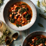 A bowl full of a tomato based Mediterranean Stew with carrots, kale, and beans throughout on top of a wooden table beside a golden spoon.