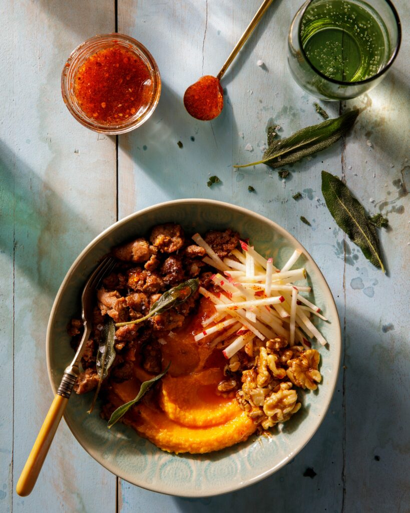 A bowl of sausage, apple sticks, butternut squash, and walnuts with a spoon in it, all laid against a blueish wood table.