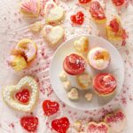 A variety of iced cookies, cupcakes and candies all in white, pink and red for Valentine's Day, on a textured white surface with white sprinkles.