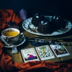 Chocolate cake served in a plate alongside tarot cards and a cup of tea