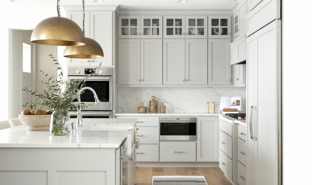 An all white kitchen depicting design trends of 2024 including cabinets and fridge with silver accents on the stove and sink and gold accents on the hooded lights above the sink.