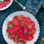 A white plate holds an array of thinly sliced beats with an herbal topping to make a Beet Carpaccio with a Citrus Vinaigrette.