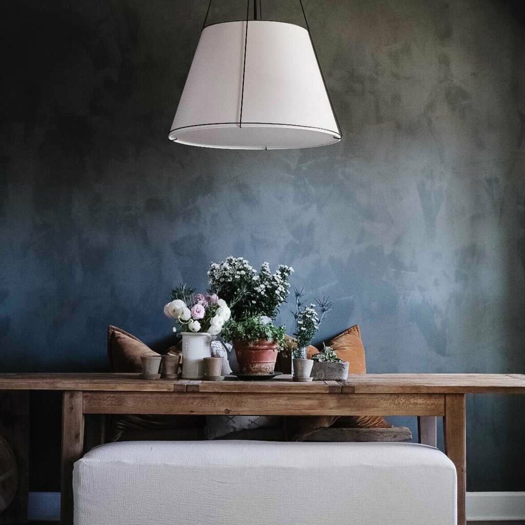 An interior shot of a dark washed wall with a wood table and grey ottoman in front of it and white light over the table.
