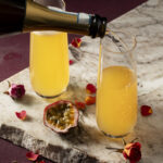 Two champagne flutes hold a yellow-orange colored drink as sparkling wine is poured into the glass on the right to make the More Adventurous Valentine's Day Cocktail.