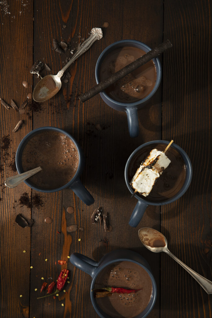 Hot Chocolate Tray with four cups of hot chocolate. One cup as a chili, one has a cylindrical chocolate bar, one has a spoon, and one has a marshmallow. hot chocolate recipes