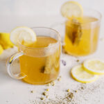 Two mugs filled with a golden hot honey toddy liquid with a tea bag in each and lemon slice on the rim. They're on a white background surrounded by spices and lemon slices.