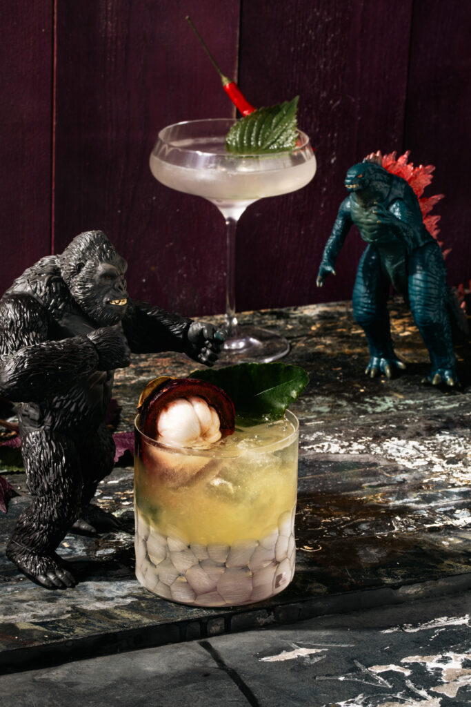 Kong and Godzilla figurines standing next to cocktails. One in a rocks glass and the other in a coupe with a deep purple background and a peeling black surface.