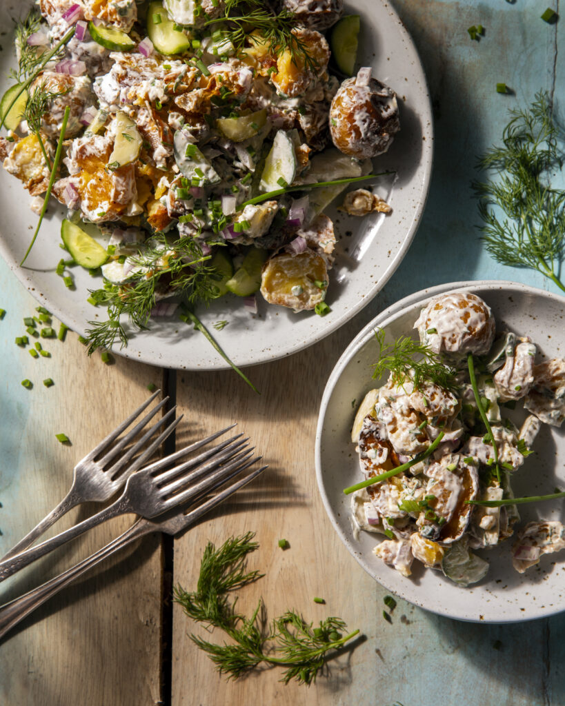 A white plate is slathered in a crispy potato salad with dill pickles and a small plate to the bottom right holds a smaller portion.