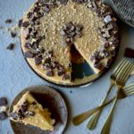 An overhead picture of a cheesecake with caramel and chocolate pieced around the edges, and a slice of the cheesecake on a plate with 3 gold forks.