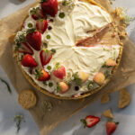 An ice cream cake pie decorated with strawberries and flowers with a slice taken out of it sits on a cutting board on a white table. Strawberries and Trefoil Girl Scout Cookies surround the pie.