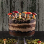 A Girl Scout Cookie Trifle layered in a clear bowl decorated with green cake moss and mushrooms on top and below the glass bowl.