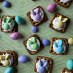 Chocolate Pretzel Easter Bunny Treats sit on a green striped background made out of pretzels, chocolate, and M&Ms with M&Ms tossed between the treats.