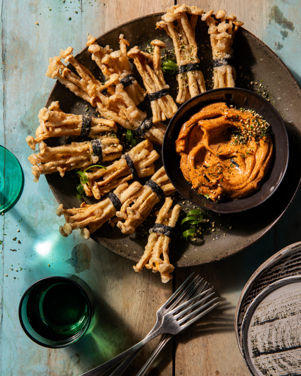 Various crispy mushroom bundles sit on a brown plate beside a small bowl of spicy mayo beside two green cups and two forks.