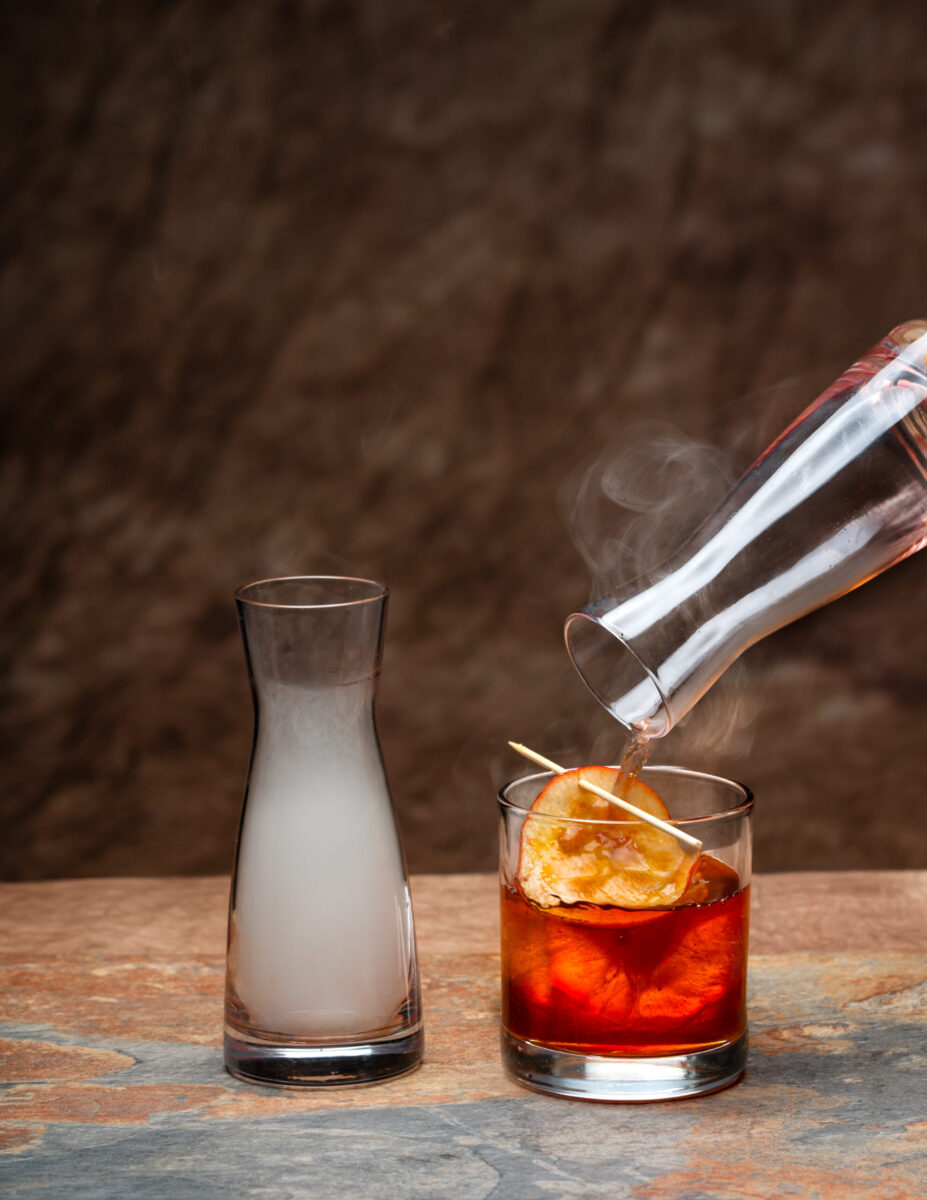 A glass filled with smoke sits beside a glass filled with an Apple Wood-Smoked Manhattan inside topped with an orange.