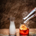 A short cocktail glass holds a red cocktail with an orange garnish next to a milk glass of smoke.