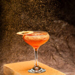 A tall cocktail glass holds an orange drink with a dried ring garnish and dusty brown sand falling overtop of it.