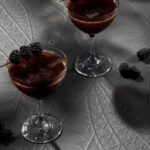 Two Back to Black cocktails in a deep brown color with a pick of blackberries on top of each glass, all on a textured grey surface.