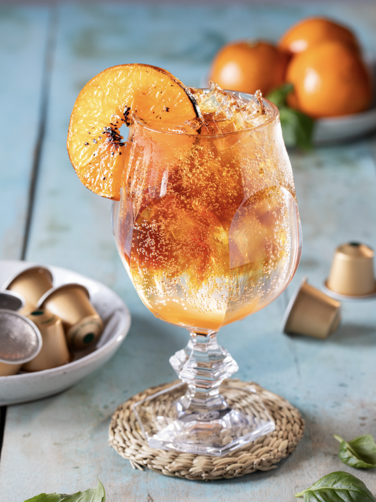 A cocktail glass is filled with a Espresso Spritz, garnished with an orange and sitting on a blue table surrounded by Nespresso pods.