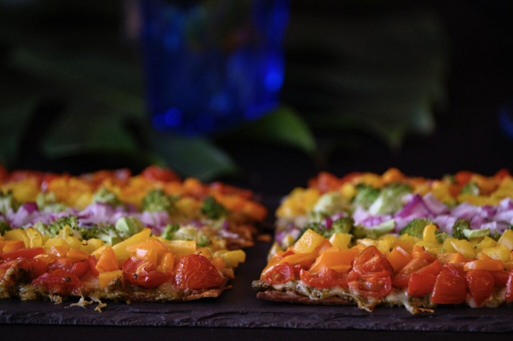 Two flatbread pizzas sitting side by side showing their thickness and rainbow veggie toppings against dark green leaves.