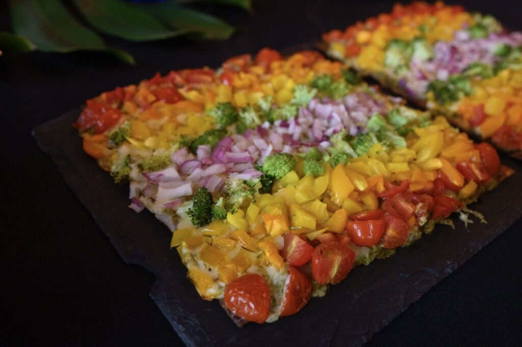 A flatbread pizza topped with veggies cut up in a rainbow design sits on a black countertop.