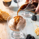 A hand pours a shot of espresso over a cup of ice cream to create a Nespresso Affogato with pizzelles and a shot of espresso in the background.