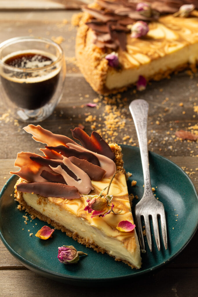 A slice of Espresso Swirl Cheesecake sits on a green plate in front of the whole baked cheesecake and a shot of espresso.