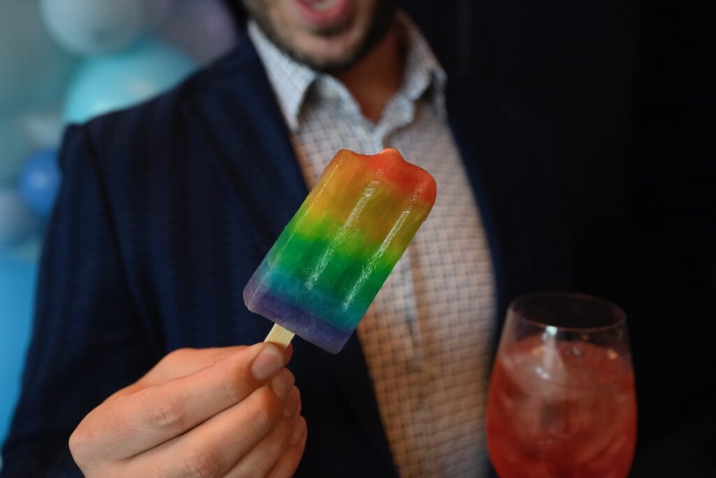 A man in a suit holds a rainbow pride popsicle in one hand with a bit taken out of it and a glass of red drink in his other hand.