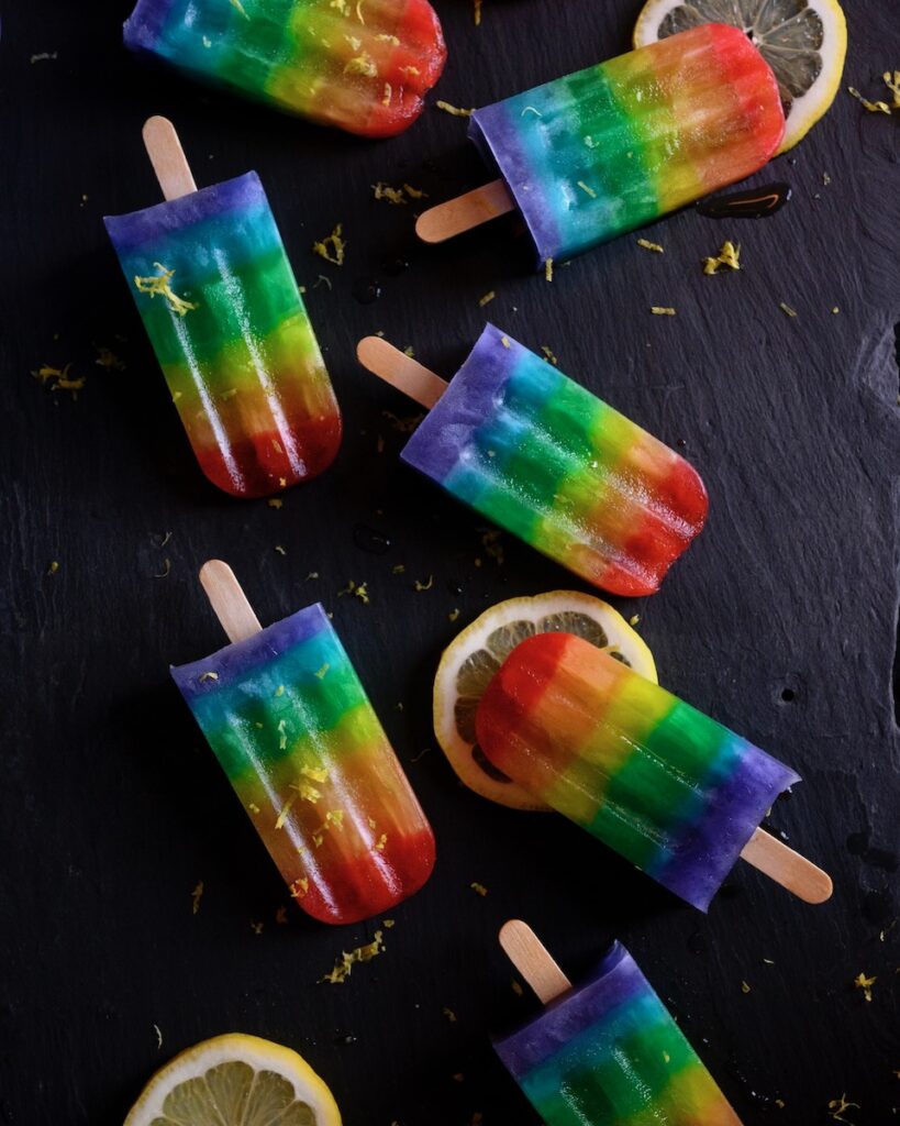 7 rainbow pride popsicles sit on a black table with slices of lemon scattered beneath them.