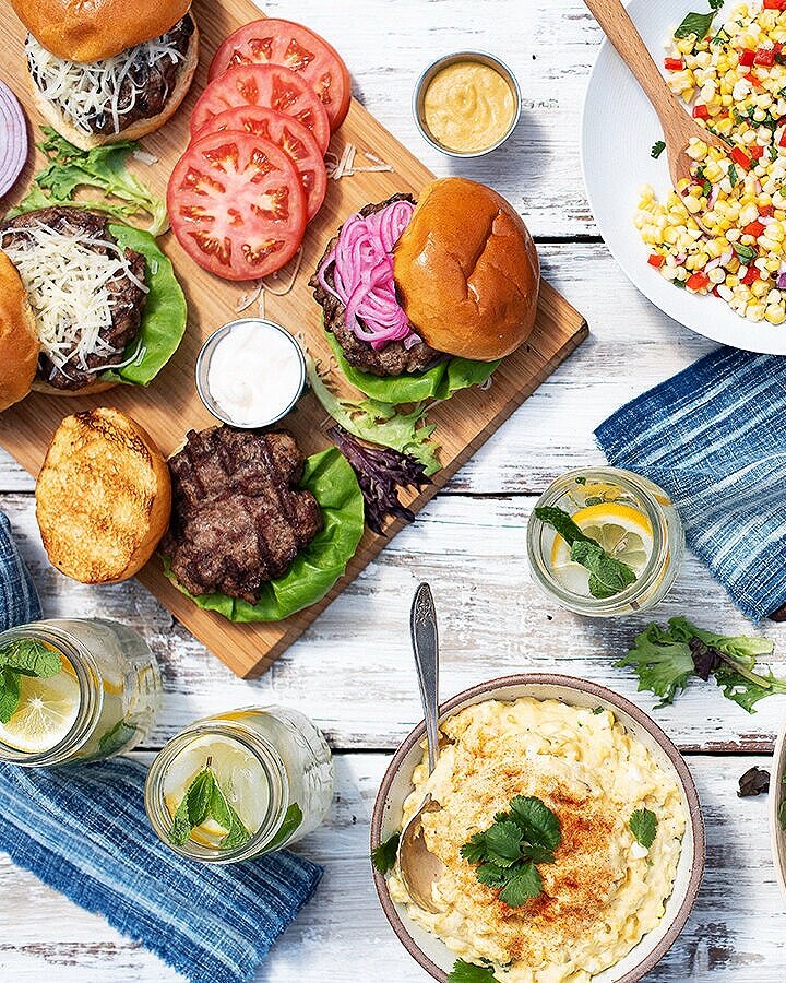 On a wood picnic table sits a wooden board covered in Great Gourmet Burgers with drinks nearby and a bowl of potato salad.