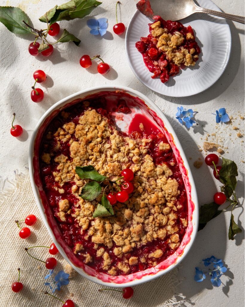 A Sour Cherry Crisp sits in a white dish with a crumble topping as a slice sits on a small white plate nearby.