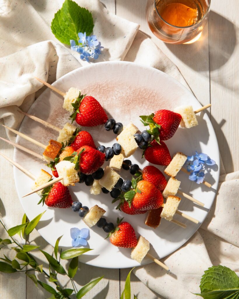 A white plate holds 6 kabobs with strawberries, blueberries, and squares of poundcake on them.