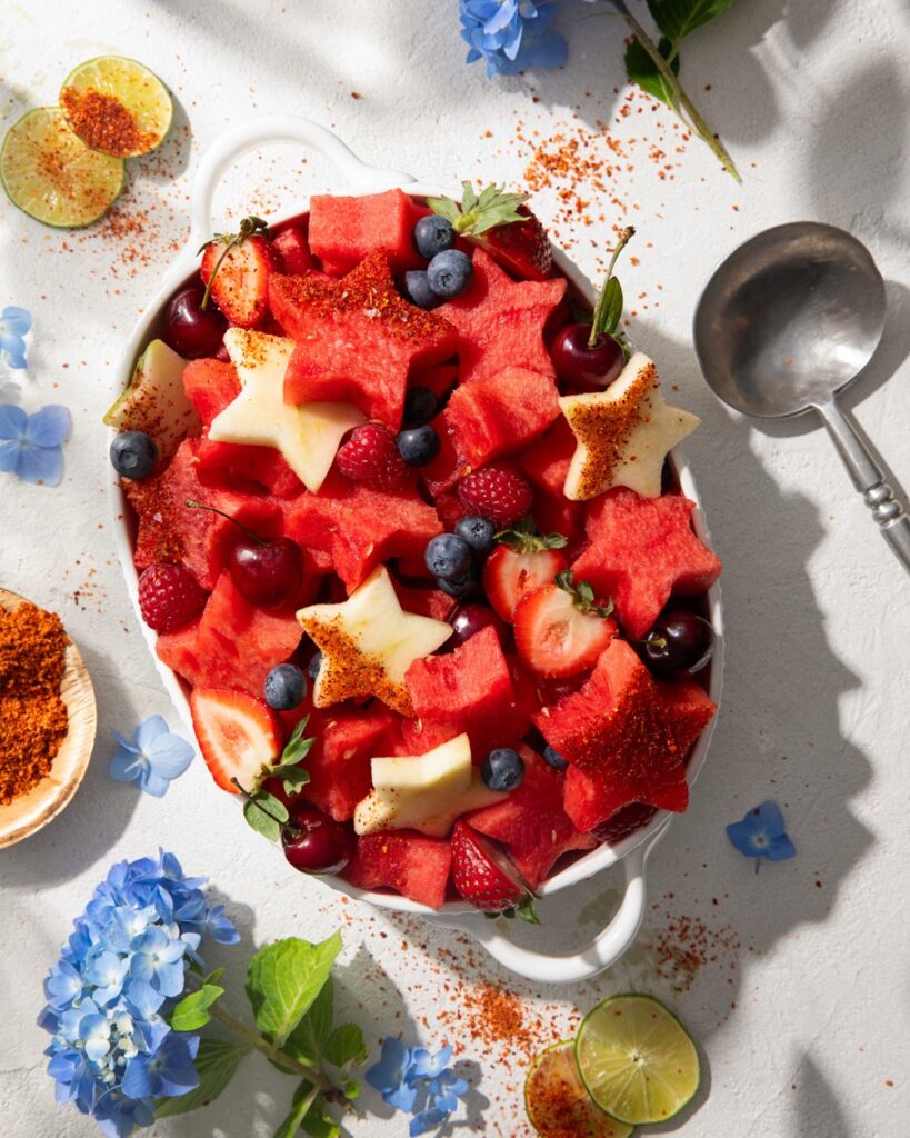 A bow sits on a picnic table filled with star shaped watermelon, strawberries, blueberries, and spinkled with tajin.