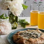 A green plate holds a greek style greens galette in front of two glasses of beer and a vase of white flowers.