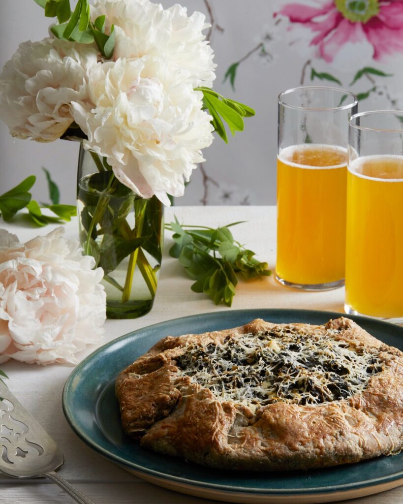 A green plate holds a greek style greens galette in front of two glasses of beer and a vase of white flowers.
