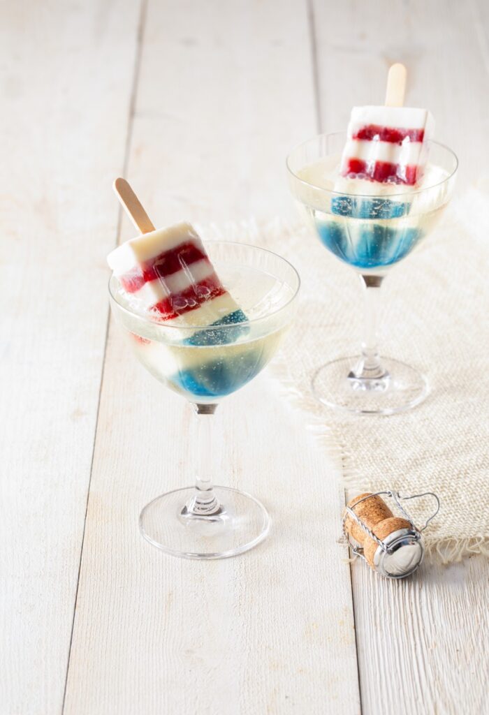 Two homemade bomb pops in red, white, and blue sit in two glasses of bubbly liquid.