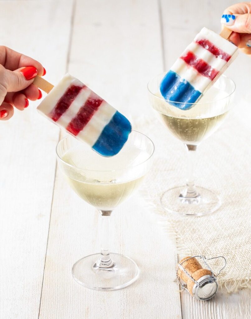 Two people hold red, white, and blue homemade bomb pops above two glasses of clear sparkling liquid on a picnic table.