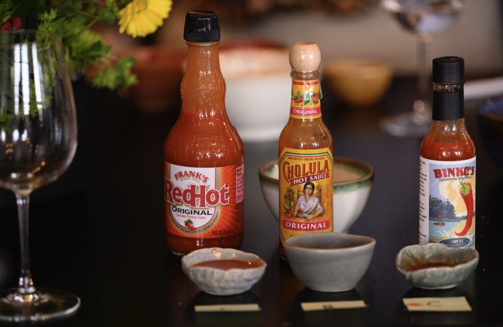 Three bottles of hot sauce sit in front of three small bowls beside a wine glass.