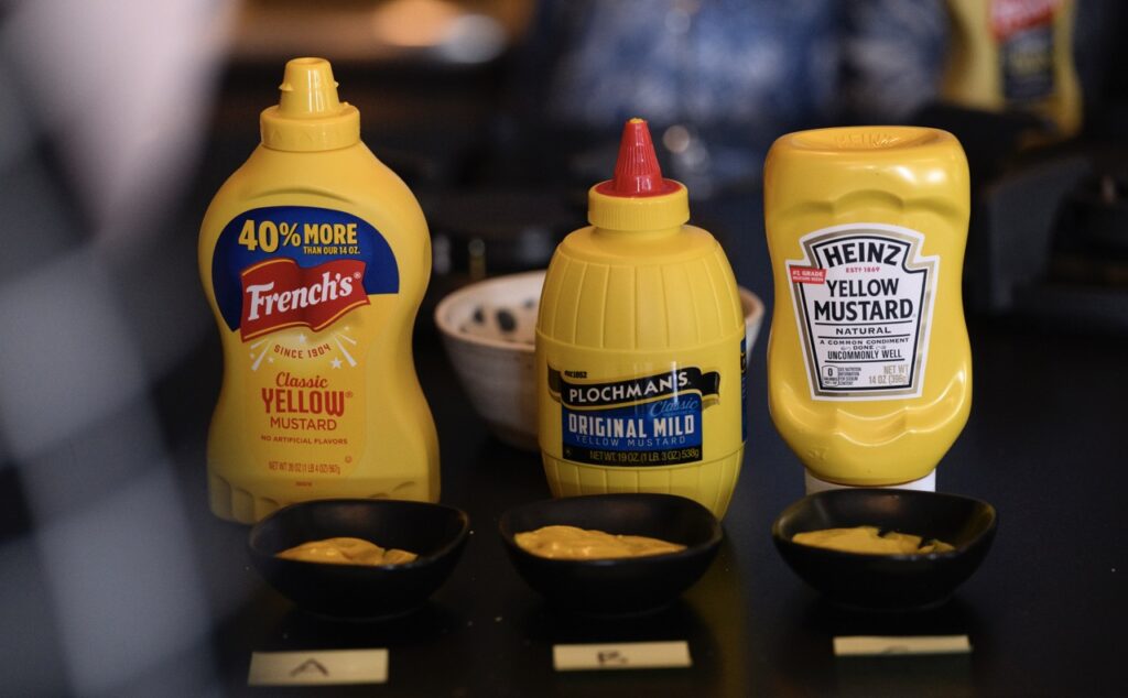 Three bottles of yellow mustard sit in front of three black containers of mustard.