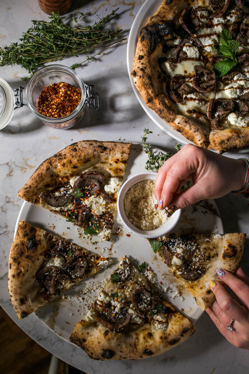 A rustic spread of charred mushroom flatbreads garnished with fresh herbs, chili oil, and a dusting of cheese, with hands reaching to sprinkle more toppings.